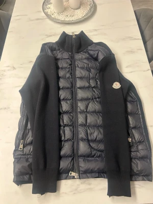 Mörkblå cardiganjacka från Moncler - Snygg mörkblå cardiganjacka från Moncler med quiltad front och stickade ärmar. Jackan har hög krage, två sidofickor med dragkedja och Moncler-logga på ärmen. Perfekt för lager-på-lager och streetstyle. Passar en M-L