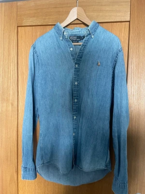 Polo Ralph Lauren, Storlek M slim fit - Säljer en avslappnad klassisk blå jeansskjorta från Polo Ralph Lauren i slim fit storlek M. Använd men i riktigt bra skick. 