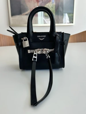 Zadig & Voltaire Candide Nono bag  - Snygg svart handväska från Zadig & Voltaire i slätt läder med silverfärgade detaljer, dragkedja och dekorativt hänglås framtill. Väskan har både handtag och en lång axelrem för olika stilar. Perfekt för dig som gillar en edgy och stilren look.