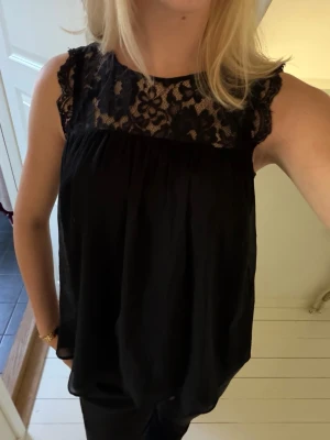 Svart ärmlös topp med spetsdetalj - Säljer en svart, luftig och ärmlös topp med vacker spets upptill vid halsen. Toppen har en lös passform och är perfekt för dig som gillar en enkel men ändå elegant stil. Materialet känns lätt och svalt, och toppen har en rund halsringning.