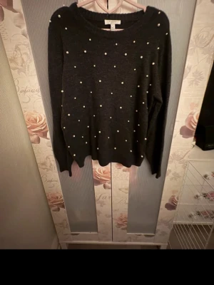 Mörkgrå stickad tröja med pärlor - Snygg mörkgrå stickad tröja med rund hals och dekorativa pärlor över hela framsidan. Tröjan har långa ärmar och ribbade muddar vid hals, ärmslut och nederkant. Perfekt för dig som vill ha en trendig och mysig look. 