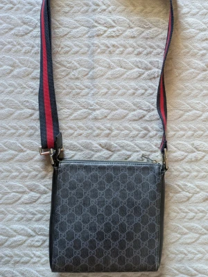 Gucci GG Supreme Small Messenger Crossbody Bag - Snygg svart axelväska med klassiskt GG-mönster och bred axelrem i rött och marinblått. Väskan har silverfärgade detaljer, dragkedja upptill och en praktisk ytterficka med dragkedja på baksidan. Perfekt för dig som gillar stilrena accessoarer.