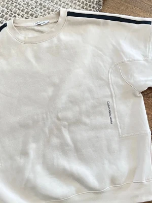 Beige sweatshirt från Calvin Klein - Beige/krämfärgad sweatshirt från Calvin Klein Jeans med svart rand längs axlarna och diskret logga på bröstet. Tröjan har rund halsringning och är tillverkad i mjukt material. Storleken är S