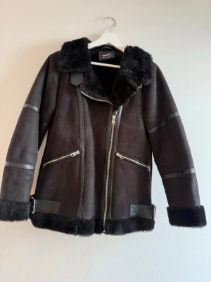Svart fuskpälsfodrad biker-kappa - Snygg svart biker-inspirerad kappa med fuskpäls vid krage, ärmslut och nederkant. Jackan har dragkedja framtill, två snedställda fickor med dragkedja och dekorativa banddetaljer. Materialet är mockaimitation och fuskpäls, perfekt för kyliga dagar. Från Pull&Bear.
