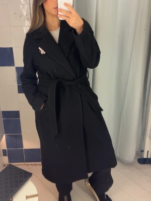 Svart lång kappa med knytskärp från vero moda - Stilren svart lång kappa från vero moda med bred krage och knytskärp i midjan. Kappan har fickor på sidorna och en enkel, klassisk design. Perfekt för kyligare dagar och ger en snygg siluett. Den är lite/medel  nopprig därav priset.