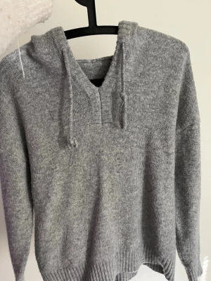 Grå stickad hoodie med huva - Mysig grå stickad hoodie med huva och dragsko. Tröjan har ribbade muddar vid ärmslut och nederkant samt en liten v-ringning framtill. Perfekt för dig som gillar en avslappnad och enkel stil. Materialet känns mjukt och värmande.