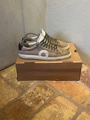 Beige sneakers med vit detalj - Snygga beige sneakers med vit sula och en stor vit cirkeldetalj på sidan. Skorna har mörkgröna skosnören och svart hälparti. Ovandelen är i mockaliknande material och insidan är vit. Perfekta för en avslappnad streetstil.