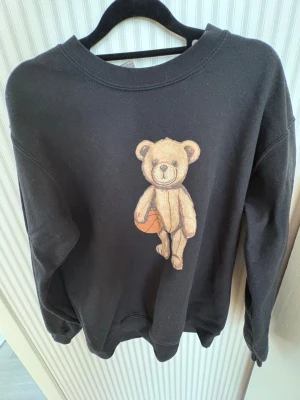 Svart sweatshirt med nalletryck - Cool svart sweatshirt med ett stort tryck av en nallebjörn som håller i en basketboll på framsidan. Tröjan har rund halsringning och ribbade muddar vid ärmslut och nederkant. Perfekt för dig som gillar streetwear och unika prints.