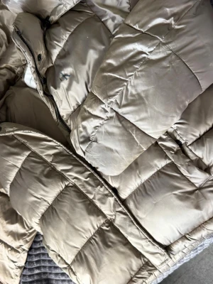 Beige pufferjacka från Calvin Klein - Snygg beige pufferjacka från Calvin Klein med diskret CK-logga på bröstet. Jackan har quiltad design, dragkedja framtill och hög krage. Perfekt för kalla vinterdagar och ger en clean och modern look.