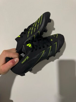 Adidas Predator svart/gul fotbollsskor  - Säljer ett par svarta Adidas Predator fotbollsskor med snygga gula detaljer och klassisk snörning. Skorna har dobbar för spel på gräs och är tillverkade i syntetmaterial. Perfekta för dig som vill ha stilrena och funktionella skor på planen.