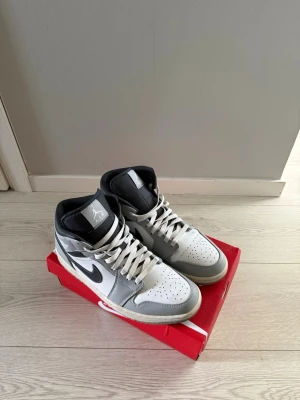 Nike Air Jordan 1 Mid grå/vit - Snygga Nike Air Jordan 1 Mid sneakers i grått, vitt och svart. Skorna har klassisk Jordan-logga på sidan, svart swoosh och vita skosnören. Ovandelen är i läder med perforerad tåbox för ventilation och vadderad krage för extra komfort.