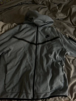Grå zip hoodie från Nike - Snygg grå hoodie från Nike med dragkedja framtill och svart Nike-logga på bröstet. Hoodien har svarta detaljer längs bröstet och ärmarna samt en stor huva. Perfekt för en avslappnad och sportig stil.