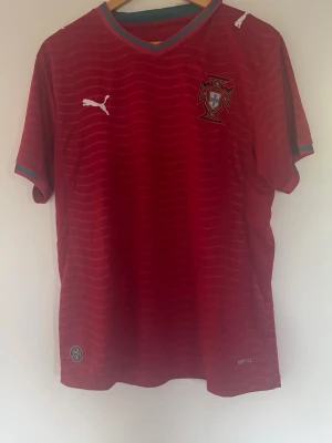 Portugal fotbollströja med shorts - Snygg röd Portugal fotbollströja från Puma med vågmönster, broderad landslagssköld och vita Puma-loggor. Tröjan har gröna och röda detaljer vid krage och ärmslut samt korta ärmar. Perfekt för match eller träning.