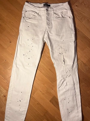 Vita Purple Jeans - Säljer ett par vita jeans från Purple. De har en diskret och snygg slitning på ena knät, och paintspalsh på både fram- och baksida. Jeansen har normal passform. Perfekta för dig som vill ha en edgy och skön look. 