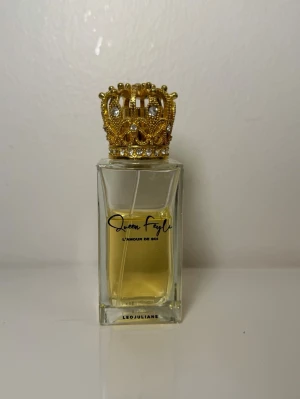 Parfym Queen Feyli  - Unik parfymflaska med guldfärgad krona som lock och genomskinlig glasflaska. Flaskan har svart text och innehåller en ljusgul vätska. Märkt med Queen Fayla och LEJULIANE. Perfekt för dig som gillar lyxiga detaljer.
