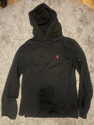 Tunn Svart hoodie från Polo Ralph Lauren - Svart hoodie med huva från Polo Ralph Lauren. Klassisk design med röd broderad logga på bröstet. Tillverkad i mjukt bomullsmaterial och har långa ärmar. Perfekt för en avslappnad och stilren look. Rätt tunn i materialet skulle mer säga att de är t shirt med luva 