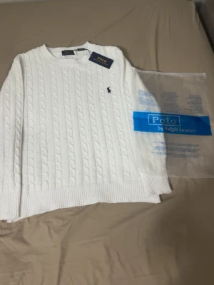 Vit kabelstickad tröja Polo Ralph Lauren - Kabelstickad vit tröja från Polo Ralph Lauren med rund halsringning och diskret broderad logga på bröstet. Tröjan har ribbade muddar vid ärmslut och nederkant, och är tillverkad i mjukt material som ger en klassisk och stilren look.