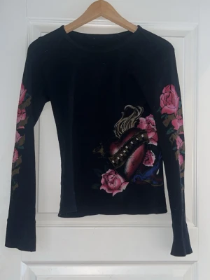 Svart Y2K-aktig tröja med blommor och hjärta i rosa, röda och blå detaljer - Svart Y2K-aktig tröja med blommor och hjärta i rosa, röda och blå detaljer. Litet hål bak på ryggen (se bild 3). Saknar märke. Jag tycker att det är en skitsnygg tröja men har knappt använt den på sistone så då är det bättre om någon annan får använda den! 🚨Tar bara emot betalningar via Swish!!! Om du vill ha fler bilder eller har frågor är det bara att skriva till mig!!!🚨