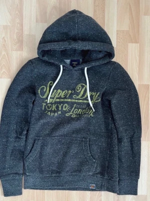 Mörkgrå Superdry hoodie med tryck - Mörkgrå hoodie från Superdry med gult tryck på bröstet där det står 'Superdry Tokyo Japan Great London Britain'. Hoodien har vit dragsko, känguruficka och mjukt material. Perfekt för en avslappnad och cool stil.