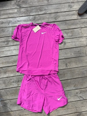 Rosa träningsset från Nike - Snyggt rosa träningsset från Nike med t-shirt och shorts. Båda plaggen har den klassiska Nike-loggan i vitt och är tillverkade i ett lätt och snabbtorkande material, perfekt för löpning eller gym. T-shirten har rund hals och korta ärmar, shortsen har resår i midjan.Passar storlek xs s
