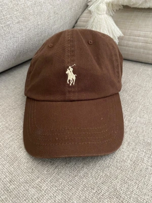 Brun keps från Polo Ralph Lauren unisex - Snygg brun keps från Polo Ralph Lauren med klassisk broderad logga framtill och texten 'POLO' baktill. Justerbar rem bak för perfekt passform. Tillverkad i bomullstyg som känns både stilrent och avslappnat.