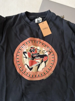 Svart sweatshirt med antikt tryck - Cool svart sweatshirt med ett stort tryck i mitten inspirerat av antik grekisk konst i rött, beige och svart. Tröjan har rund hals och långa ärmar. Perfekt för dig som gillar unika och konstnärliga plagg.