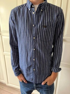 Blåvit randig skjorta från Lee - Snygg blå och vit randig skjorta från Lee med klassisk krage och knappstängning framtill. Skjortan har en bröstficka med Lee-logga och långa ärmar. Perfekt till jeans för en avslappnad men stilren look. Skjortan är i storlek M. Hör av er vid frågor och priset är inte hugget i sten!