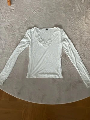 Vit långärmad topp med spets - Säljer en vit långärmad topp med v-ringning och fina spetsdetaljer runt halsen och runt wristen. Toppen har en slim passform och är tillverkad i ett mjukt material som känns skönt mot huden. Perfekt att styla med jeans eller kjol.