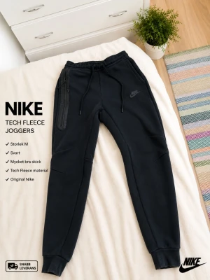 Svarta Tech Fleece mjukisbyxor Nike - Säljer ett par svarta Tech Fleece mjukisbyxor från Nike. Byxorna har dragsko i midjan, ribbade muddar vid bensluten och en snygg ficka med dragkedja på sidan. Perfekta för chill eller träning, riktigt sköna och stilrena.