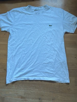 Vit t-shirt från Lacoste - Klassisk vit t-shirt från Lacoste med den ikoniska gröna krokodilloggan broderad på bröstet. T-shirten har rund hals och korta ärmar, tillverkad i mjuk bomull för en skön och enkel stil.