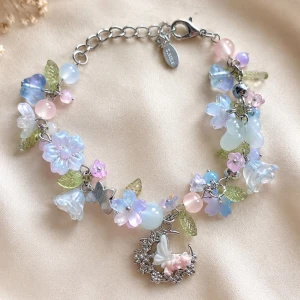Pastellfärgat armband med blommor - Säljer ett armband med pärlor och blommor i pastellfärger som rosa, blått och lila. Armbandet har små gröna blad och en söt silverfärgad berlock med en måne och en fjäril. Perfekt för dig som gillar gulliga och unika accessoarer. Längd 22cm kan justeras mellan 16-21cm 
