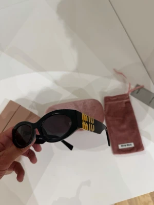 Svarta solglasögon från Miu Miu 11w - Stilrena svarta solglasögon från Miu Miu med bred båge och diskret gulddetalj vid skalmen. Kommer med original rosa sammetsfodral och putsduk. Perfekta för dig som vill ha en lyxig accessoar med statement-känsla.