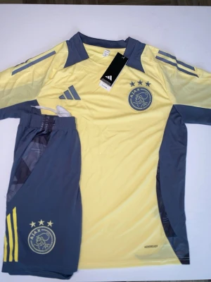 Ajax träningsset från Adidas gul/grå - Ajax träningsset från Adidas i gul och grå färg. Setet består av en kortärmad tröja med Ajax-logga och Adidas-ränder samt matchande shorts med tryckt klubbmärke och gula detaljer. Tillverkat i lätt och ventilerande material, perfekt för fotbollsträning.
