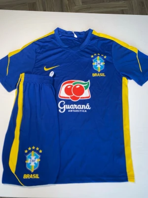 Blått Brasilien fotbollsställ - Säljer ett blått Brasilien fotbollsställ med gula detaljer. Setet består av tröja och shorts, båda med CBF-logga och sponsortryck. Materialet är lätt och andas, perfekt för träning eller match. Snygg design med gula ränder längs sidorna och tryck på både fram- och baksida.