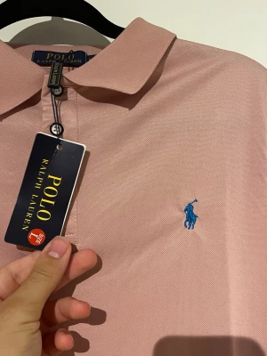 Ljusrosa pikétröja från Polo Ralph Lauren - Säljer en ljusrosa pikétröja från Polo Ralph Lauren med klassisk krage och blå broderad logga på bröstet. Tröjan har korta ärmar och knappar vid halsen. Perfekt för en clean och stilren look.
