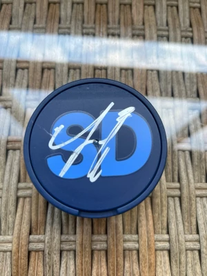 SD dosa - signerad av Jimmie Åkesson - En rund popsocket i svart plast med blå SD-logga och vit signatur på ovansidan. Perfekt som grepp eller stöd till mobilen och enkel att fästa på baksidan av telefonen.