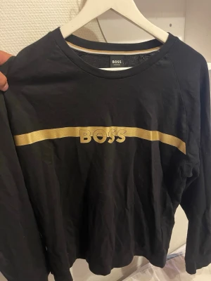 Svart långärmad tröja från BOSS - Snygg svart långärmad tröja från BOSS med guldfärgad logga och rand över bröstet. Tröjan har rund halsringning och är tillverkad i mjukt bomullsmaterial. Perfekt för en stilren och avslappnad look. Skriv om du har frågor om tröjan eller vill ha mer bilder