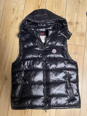 Svart dunväst från Moncler med huva - Snygg svart dunväst från Moncler med glansig finish, avtagbar huva och två dragkedjeförsedda fickor. Klassisk Moncler-logga på bröstet och quiltad design. Perfekt för lager på lager och streetwear-vibe.