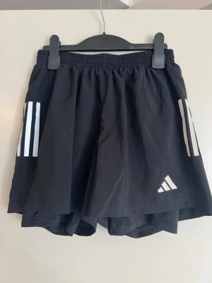 Svarta Adidas sportshorts - Svarta sportshorts från Adidas med vita ränder på sidorna och Adidas-logga framtill. Shortsen har elastisk midja och innerbyxa för extra stöd. Tillverkade i lätt och snabbtorkande syntetmaterial, perfekta för träning och aktivitet. Köpta på herr men funkar även på dam, jag som tjej har använt dem.   Regular fit, har en bakficka med dragkedja och innerbyxa. Inköpta på Stadium. Använda 2 gånger.   Midja: ca 32cm Längd: ca 34cm