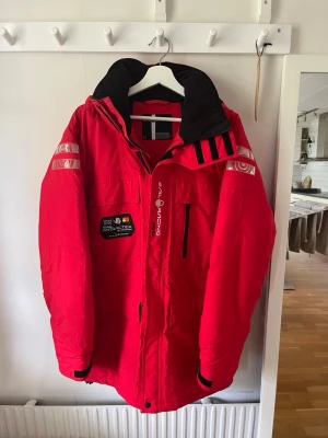 Röd parkas med reflexdetaljer Sail racing - Röd parkas med svart huva och reflexdetaljer på ärmarna. Jackan har flera fickor, dragkedja framtill och patch med texten 'Antarctica' och 'Sail Racing' på bröstet. Perfekt för kalla dagar och outdoor-äventyr.