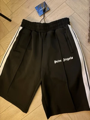 Palm Angels shorts  Nya med tags -   Säljer ett par Palm Angels shorts i svart med vita sidoränder och vit logga. Helt nya med tags kvar. Bekväm passform med elastisk midja och fickor med dragkedja. Perfekta till både vardag och sommar. 
