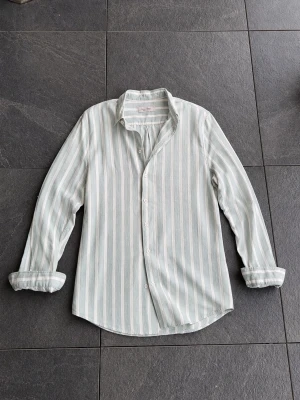 Randig linneskjorta från Dressmann - Säljer en ljusblå och vit randig skjorta från Dressmann i mjukt linne. Skjortan har klassisk krage, långa ärmar och knappar framtill. Perfekt för dig som gillar en avslappnad men stilren look.