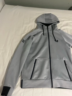 Under aurmor hoodie med dragkedja  - Snygg ljusgrå hoodie från Under Armour med hel dragkedja framtill, svarta detaljer och två fickor med dragkedja. Justerbar huva med snören och logga på bröstet samt ärmen. Perfekt för en sportig och avslappnad stil.