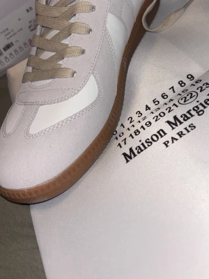 Maison Margiela vita sneakers - Maison Margiela sneakers i vitt och beige med klassisk gummisula. Skorna har en stilren design med mockadetaljer och vita läderpaneler. Beige snörning och låg profil ger en clean och modern look. Perfekta för dig som gillar minimalistisk stil.