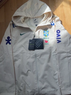 Vit Brasil Nike vindjacka med huva - Snygg vit vindjacka från Nike med Brasiliens landslagslogga och flera tryck i blått och grönt. Jackan har huva, hel dragkedja och är tillverkad i ett lätt, tekniskt material. Perfekt för dig som gillar sportig stil och vill sticka ut.