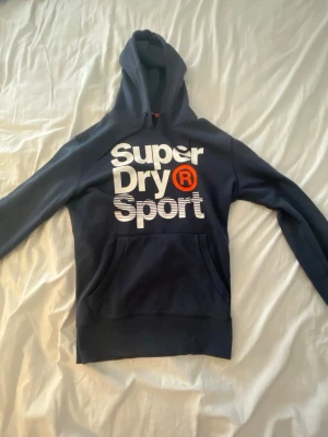 Mörkblå Superdry Sport hoodie - Snygg mörkblå hoodie från Superdry Sport med stor vit logga och röd detalj på bröstet. Klassisk huva med dragsko och känguruficka framtill. Perfekt för en sportig och avslappnad stil.