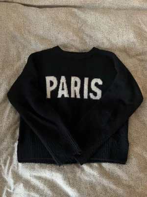 Svart stickad tröja med PARIS-tryck - Mysig svart stickad tröja med vit text 'PARIS' framtill. Tröjan har rund halsringning, långa ärmar och ribbade muddar vid ärmslut. tyvärr för kort för mig 🖤🤍