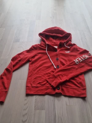 Röd hoodie från Hollister med tryck - Röd hoodie från Hollister med knäppning framtill, vit dragsko i huvan och tryckt logga på ena ärmen. Klassisk Hollister-fågel på bröstet. Mjuk bomullskänsla och långa ärmar. Med dem eftertraktade knapparna passar den perfekt för en avslappnad streetstil.