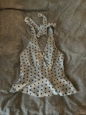 Prickig vit omlottopp med knyt - Snygg vit topp med svarta prickar och omlottdesign. Toppen har djup v-ringning, bred midja och volangkant nedtill. Knytband i nacken ger en trendig look och toppen är ärmlös. köpte den på plick !! 🖤🤍