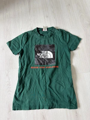 Grön t-shirt från The North Face - Snygg grön t-shirt från The North Face med klassisk logga och texten 'Never Stop Exploring' i rött framtill. T-shirten har rund hals och korta ärmar, perfekt för en avslappnad och sportig stil.